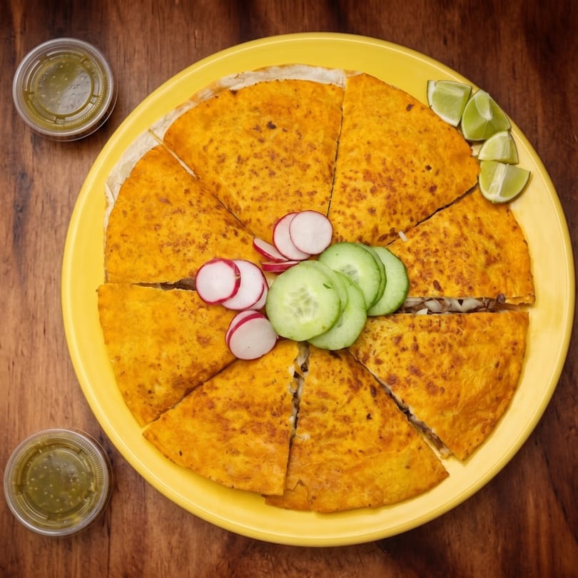 A large, golden-brown quesadilla cut into eight slices, garnished with radishes, cucumbers, and lime wedges.