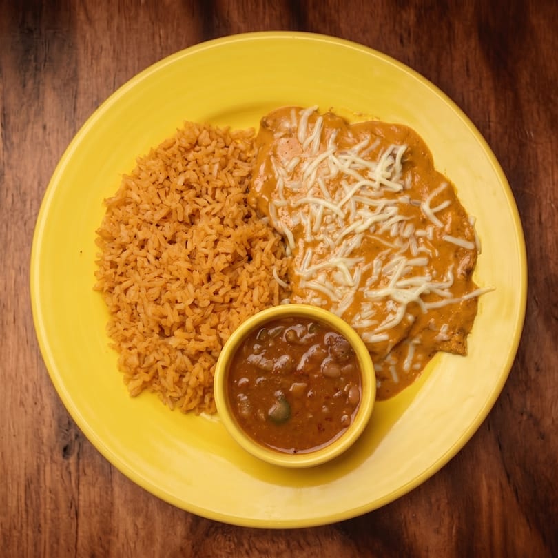 A yellow plate holds two enchiladas with orange sauce and shredded cheese, rice, and a side of beans.