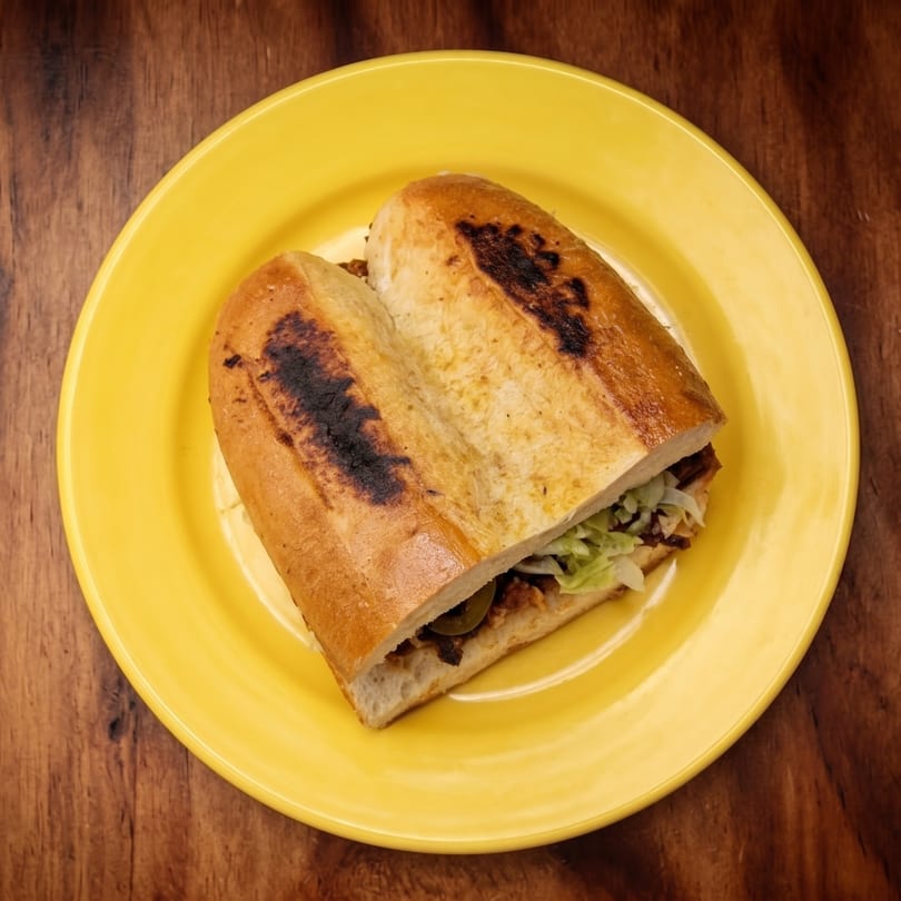 A toasted torta sandwich, cut in half, filled with meat, lettuce, and jalapeños on a yellow plate.