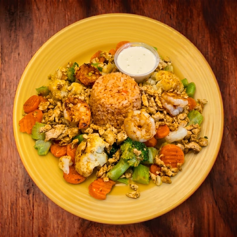 A yellow plate holds a colorful meal of rice, shrimp, chicken, broccoli, carrots, and cauliflower with a side of white sauce.